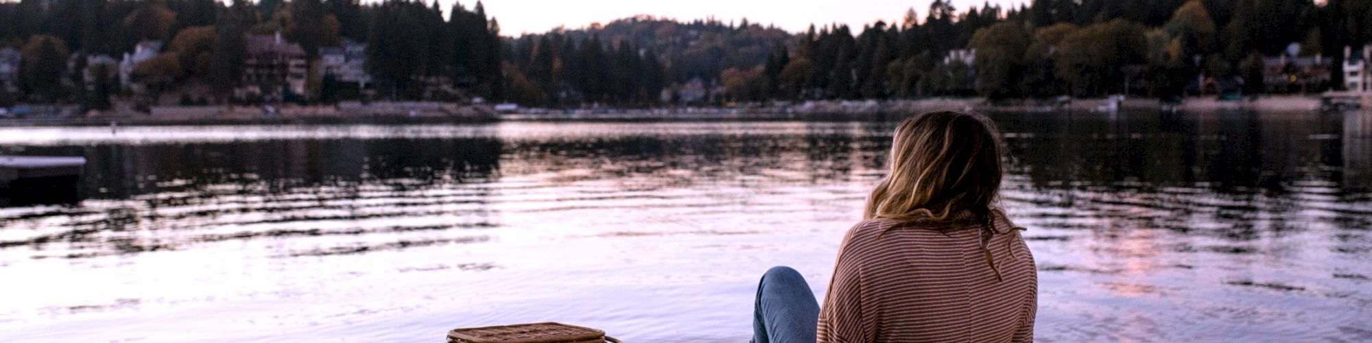 A person sits by a lake at dusk, a picnic basket nearby.