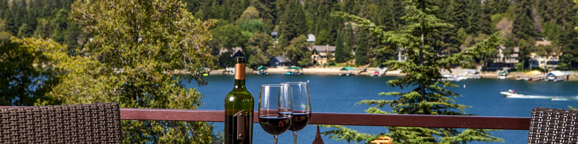 Wine bottle, glasses, and food sitting on top of a table looking over a beautiful lake view with trees in the background..