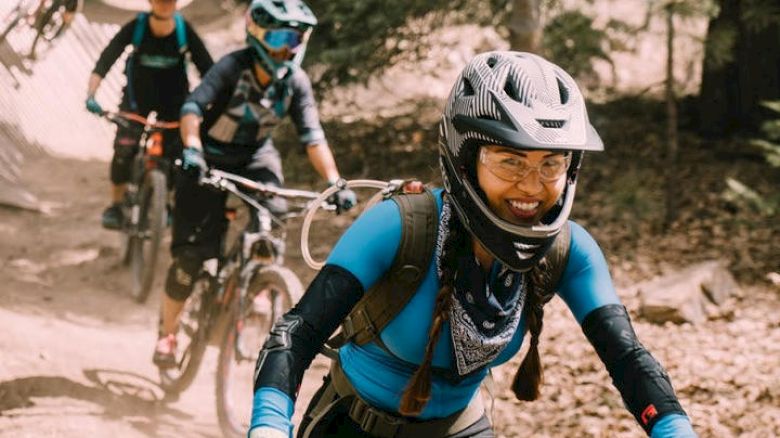 Cyclists with helmets ride mountain bikes on a dirt trail in a forested area, wearing sporty gear and backpacks, one smiling at the camera.