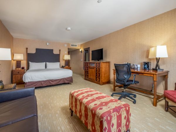 A hotel room with a bed, desk, chair, TV, sofa, and a padded bench. Lamps provide lighting, and the decor is warm and inviting.