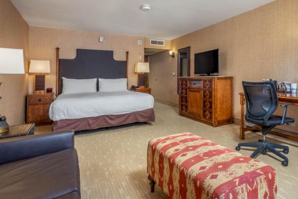 A hotel room with a large bed, patterned bench, desk with chair, and carpeted floor.