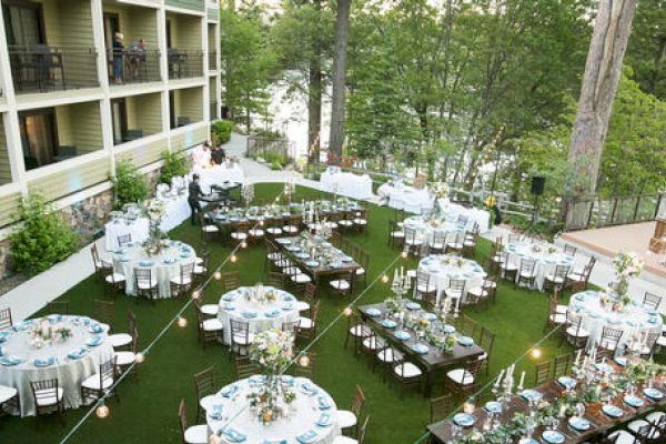 Outdoor event setup with tables and chairs on a lawn, surrounded by trees and a building.