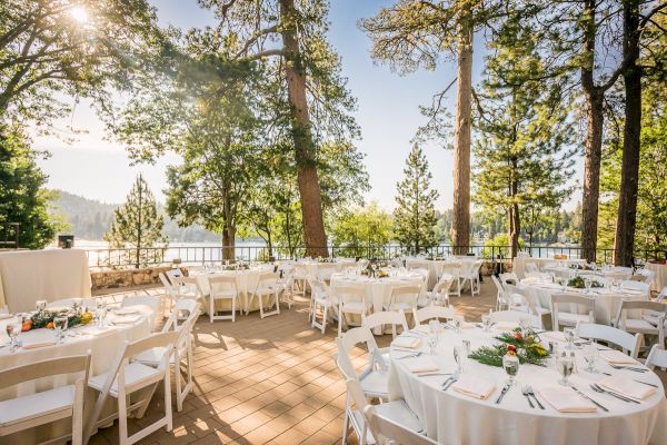 Outdoor event setup with white tables and chairs among trees with a lake view.