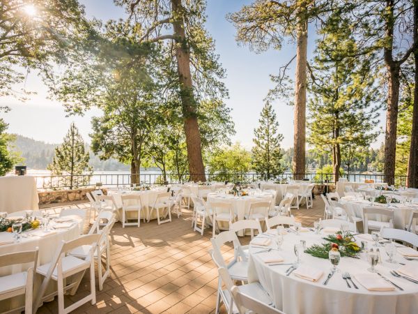Outdoor event setup with white tables and chairs among trees with a lake view.