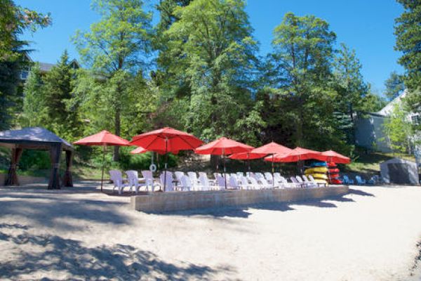 Sandy beach with red umbrellas, white chairs, and a gazebo amidst green trees.