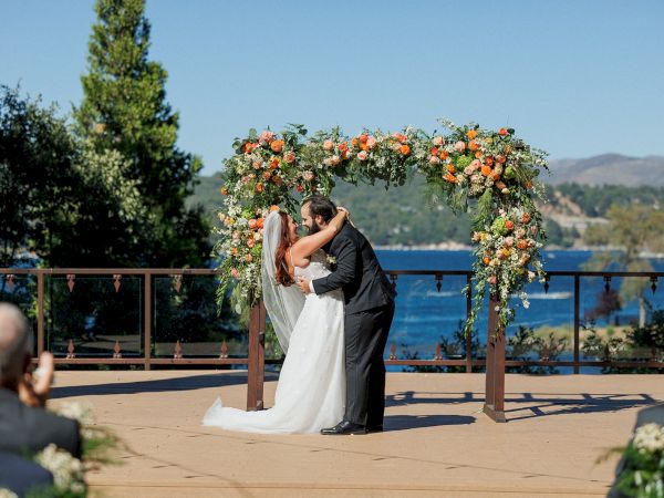 A couple is kissing under a floral arch at an outdoor wedding ceremony with a scenic background of trees and a body of water.