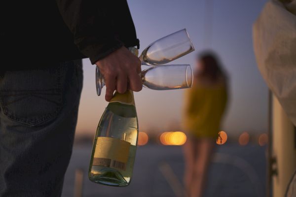 Two people walking toward a beach at nighttime, one of which is holding a bottle of sparkling wine and two champagne flutes.
