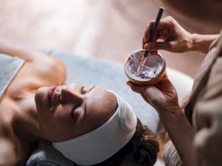A person is lying down for a facial treatment with a towel around, while another applies a facial mask using a brush from a small bowl.