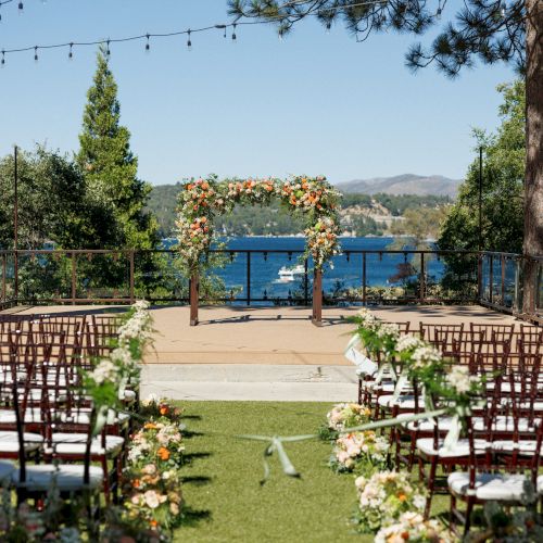 An outdoor wedding setup with an aisle, chairs, floral arrangements, a decorated arch, and a scenic view of a lake and trees.