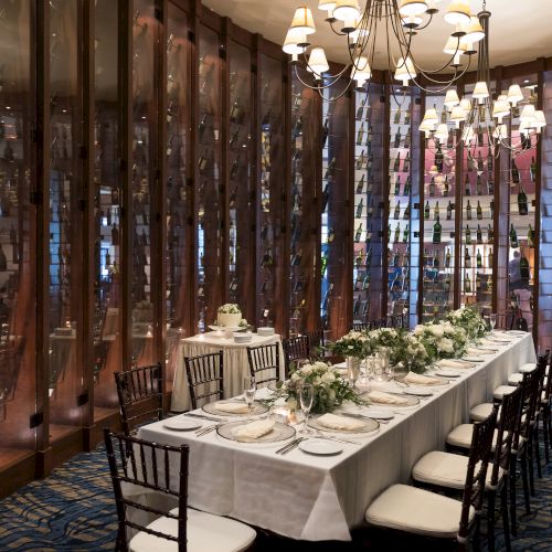 A long table is elegantly set for a formal dinner, surrounded by wine racks and soft lighting from chandeliers in a luxurious room.