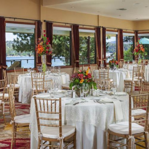 A decorated banquet hall with round tables, white tablecloths, chairs, and floral centerpieces, overlooking a scenic outdoor view.
