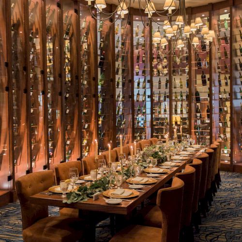 A long dining table set for a meal in an elegant room with warm lighting and wine racks lining the walls, creating an upscale ambiance.