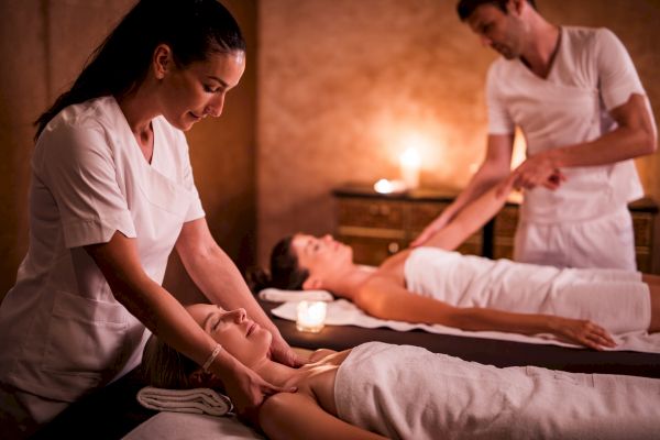 Two people receiving massages in a dimly lit spa, with massage therapists attending to them, enhancing relaxation and well-being.