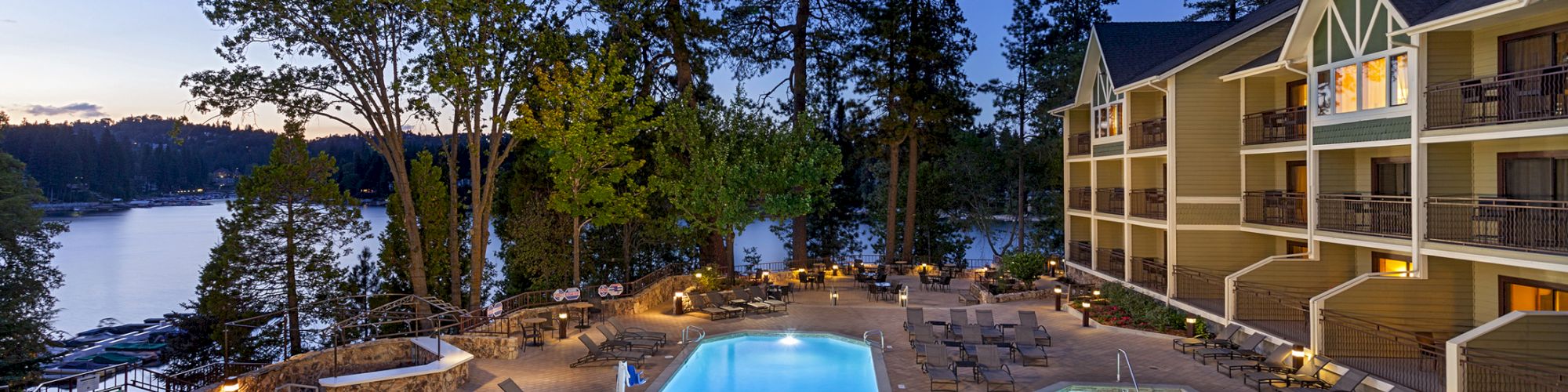 A serene hotel pool area at dusk with lounge chairs and trees, overlooking a lake and surrounded by a multi-story building.