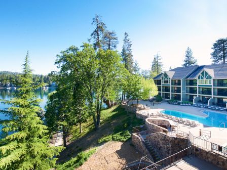 A scenic view of a lakeside resort features a building, a pool, a hot tub, and lush trees surrounding a calm lake under a clear sky.