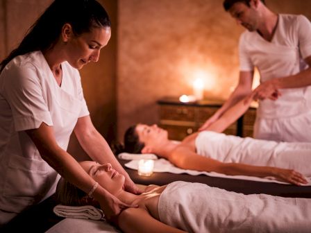 Two people receive massages on spa beds, attended by therapists in a calming room with candles and a warm ambiance.