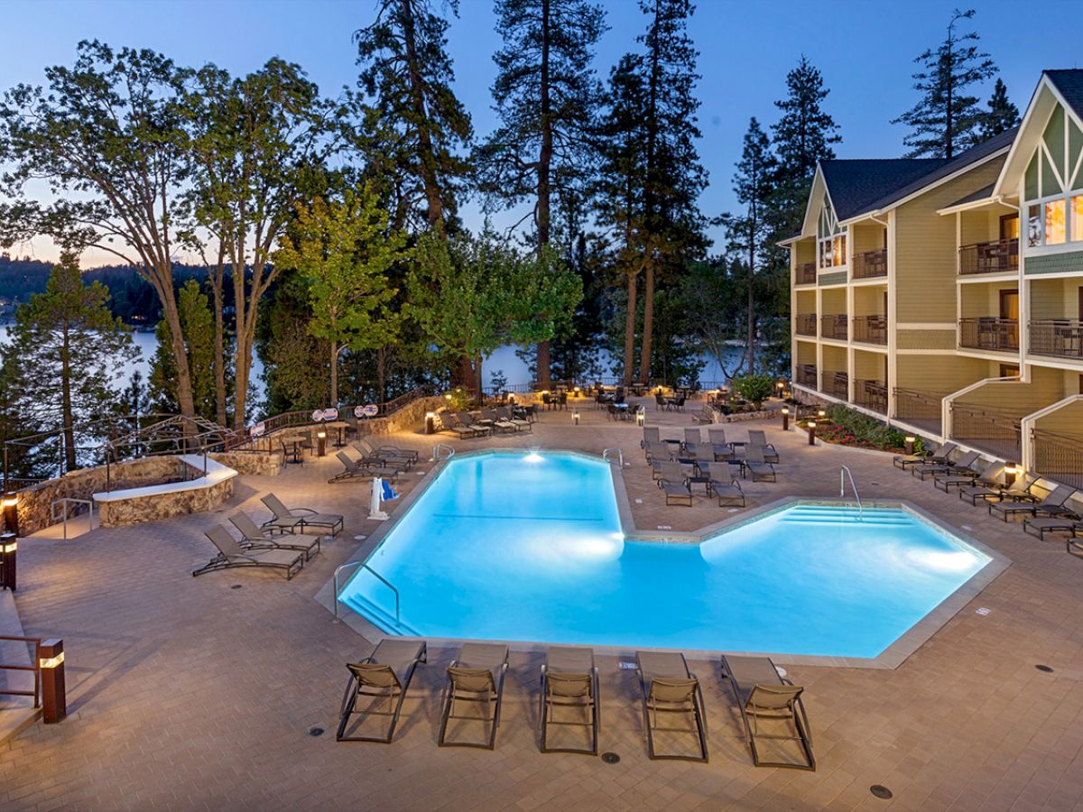 A poolside area with lounge chairs, near a building by a lake surrounded by trees, captured at dusk.