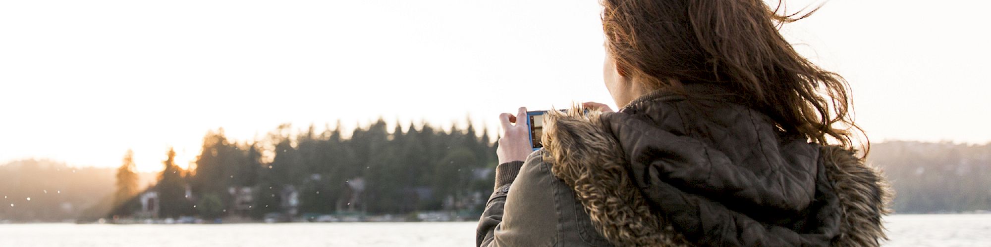 A person in a jacket, on a boat, takes a photo of water and a tree-lined shore at sunset.