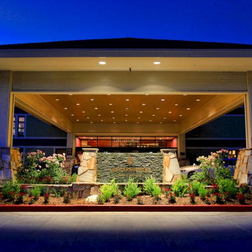 The image shows the entrance of a building with a covered driveway, stone pillars, and landscaped gardens at night.