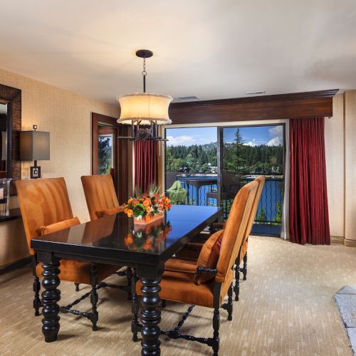 A dining room with a table, orange chairs, a floral centerpiece, stone fireplace, and a scenic view through large windows with red curtains.