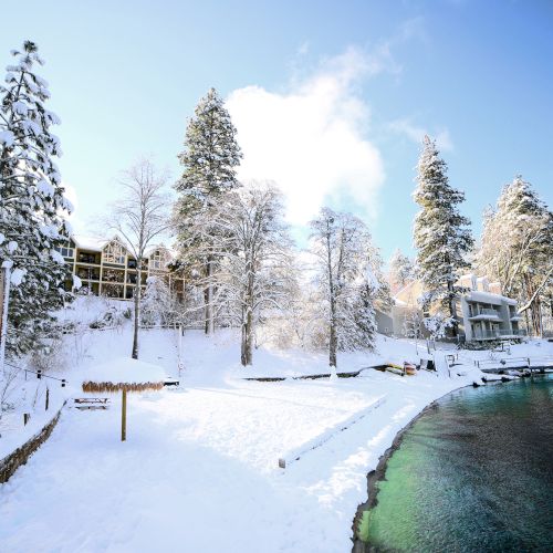 A snowy landscape with trees and buildings by a clear, serene body of water under a bright blue sky.
