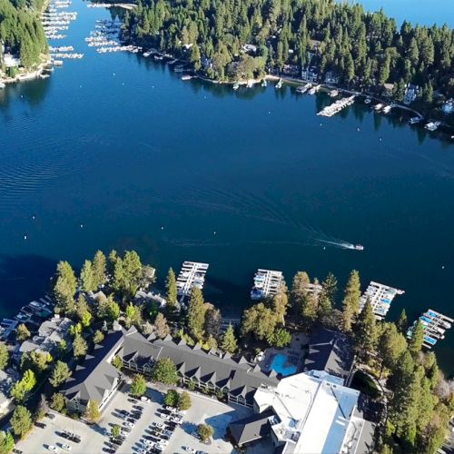 Aerial view of a lake surrounded by forest, docks with boats, and buildings with a parking lot in the foreground.
