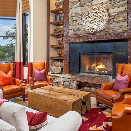 Cozy living room with leather chairs, stone fireplace, wood table, colorful cushions, decorative shelves, large windows, and vibrant drapes.
