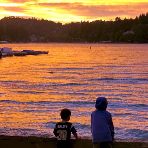 Two children stand by a lakeside during a vibrant sunset, with boats docked nearby and a tree-lined horizon.