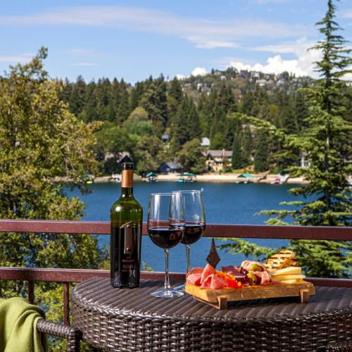 A table with wine, glasses, and a charcuterie board overlooks a scenic lake surrounded by trees and hills under a clear sky.