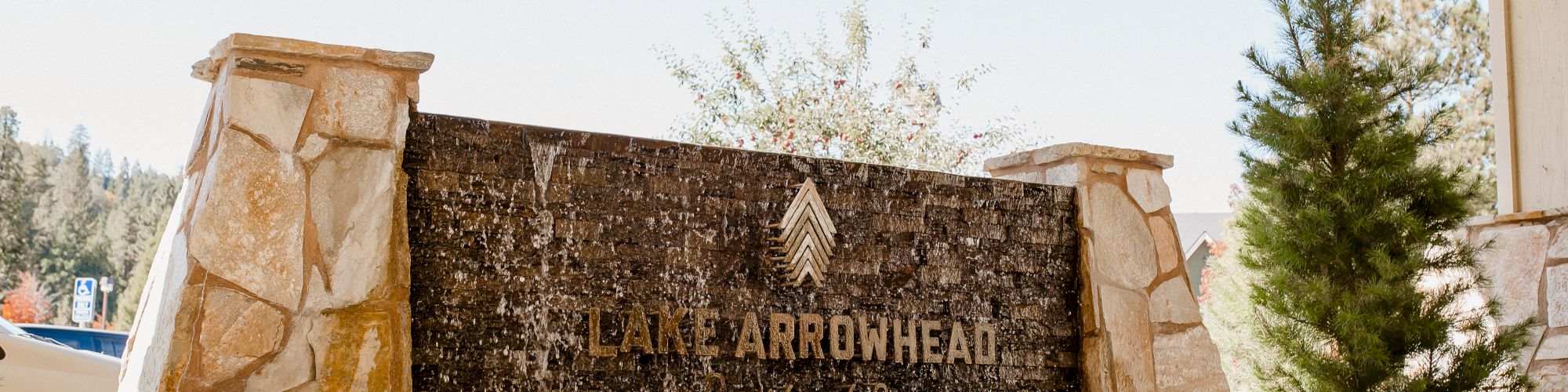 The image shows a sign for "Lake Arrowhead" surrounded by shrubs, stone pillars, and trees, beneath a structure with a canopy overhead.