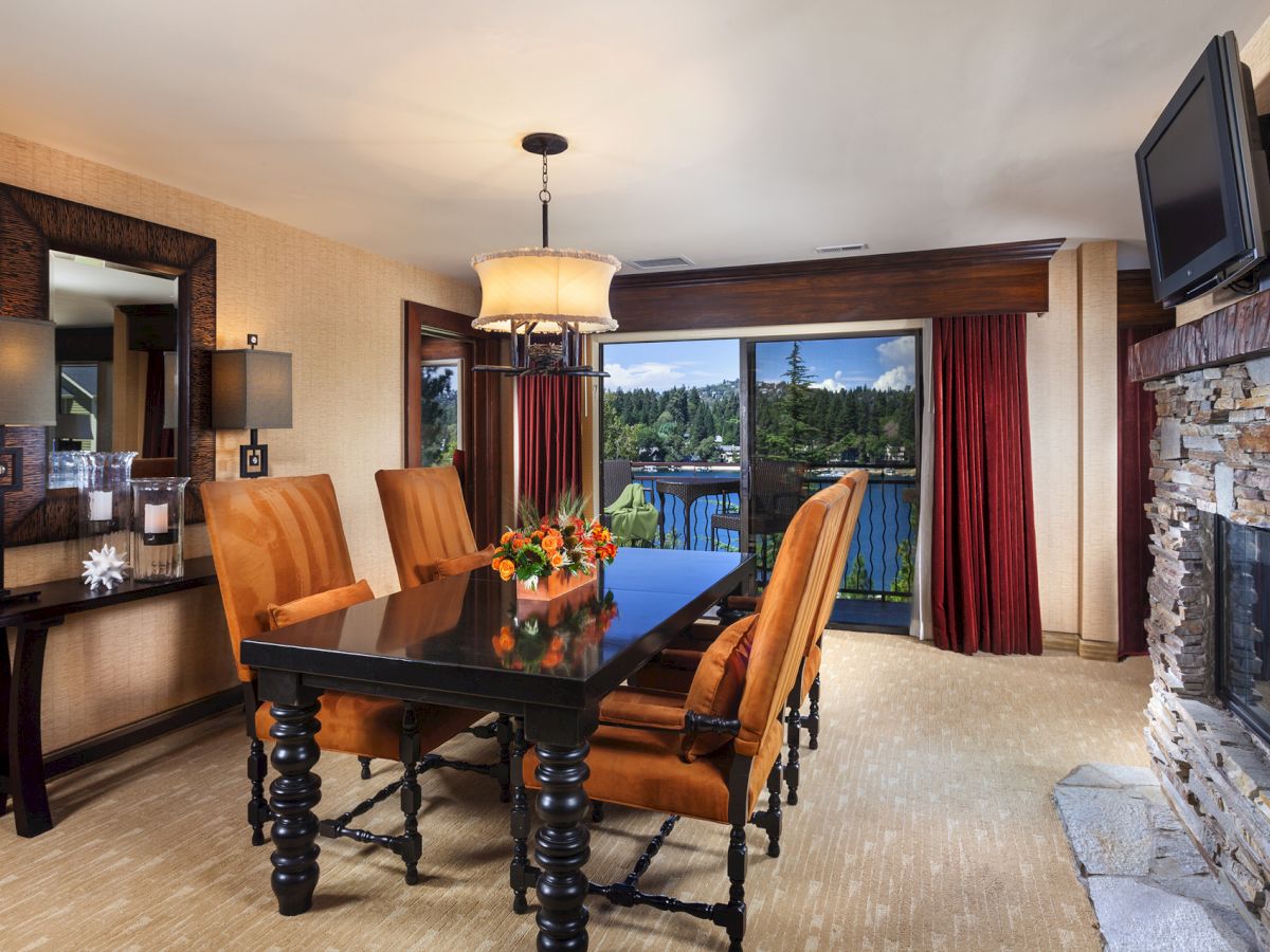 A dining room features a wooden table, orange chairs, a large mirror, fireplace, and a window view of trees and a lake.