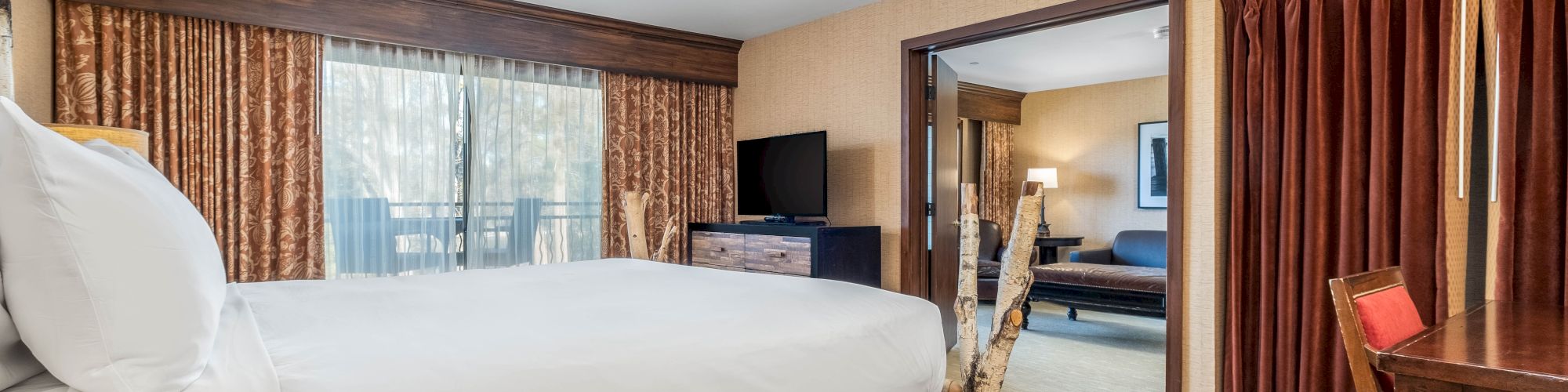 A hotel room with a bed, curtains, TV, and a desk, featuring an adjoining seating area with a couch and chair.