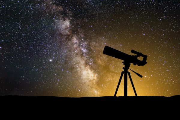 A silhouette of a telescope stands against the starry sky, with the Milky Way galaxy visible in the background.