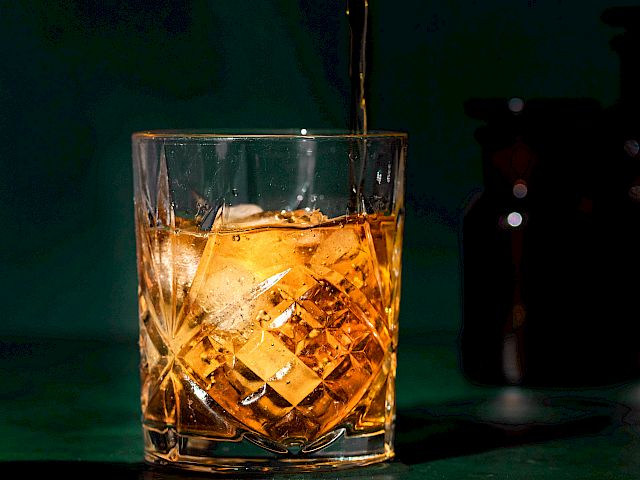 A glass with ice is being filled with a golden liquid, possibly whiskey or similar beverage, against a dark background.