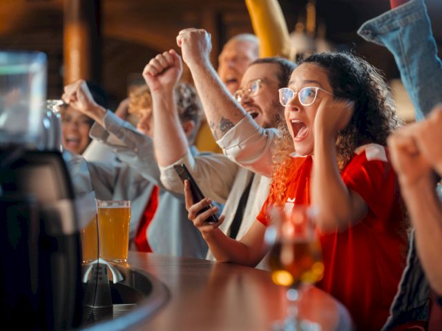 A group of excited people at a bar, cheering and raising their fists, possibly watching a sports event on TV, with drinks in hand.