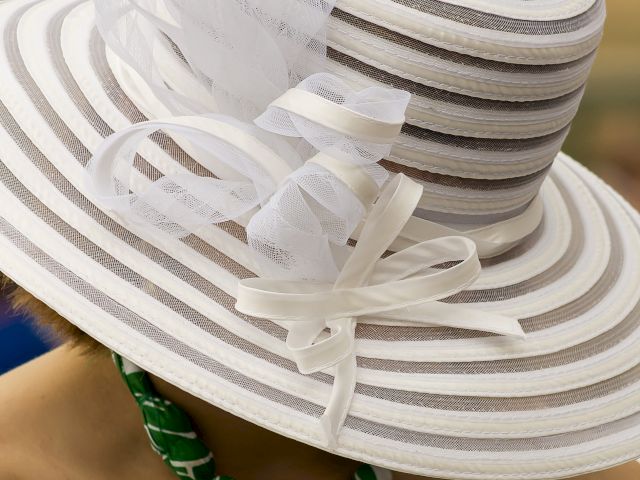 A person wearing a wide-brimmed, striped hat with decorative white ribbons and a green beaded necklace.