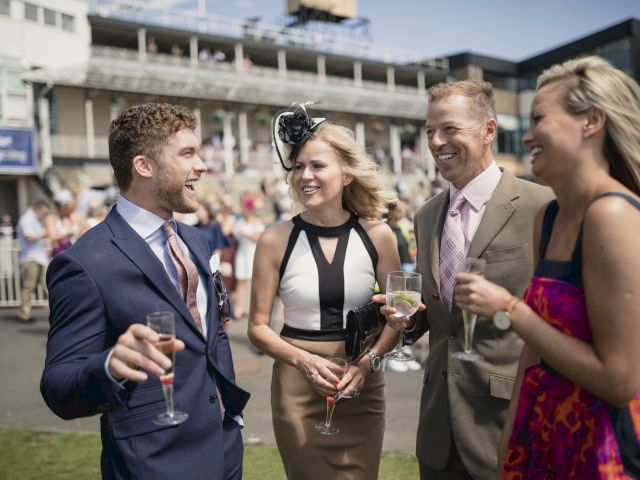 A group of four people dressed formally, laughing and holding drinks, outdoors at an event with a building in the background.