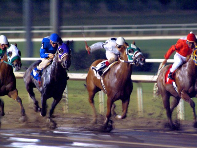 Jockeys compete in a dynamic horse race on a muddy track, each wearing distinct colors under the stadium lights, creating an intense scene.