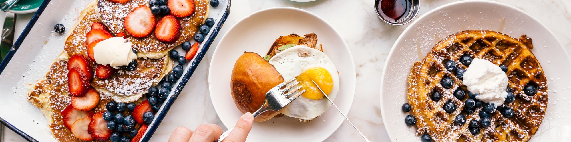 Breakfast spread with pancakes, strawberries, blueberries, a waffle, egg sandwich, coffee, and utensils on a white tabletop.