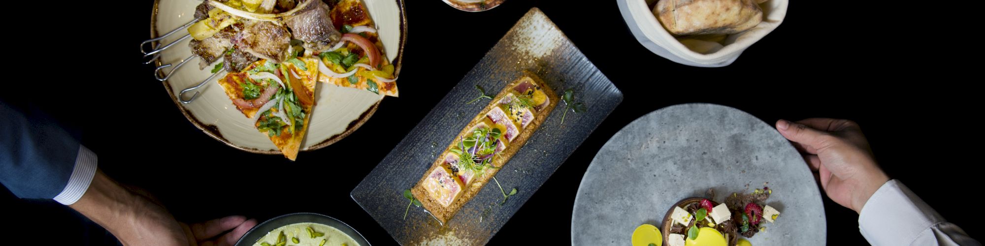 A variety of dishes are presented on a black background: pizza, bread, fish, salad, and desserts, with hands reaching for them.