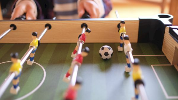 The image shows a foosball table with players in yellow and red uniforms, a ball in the center, and someone playing.