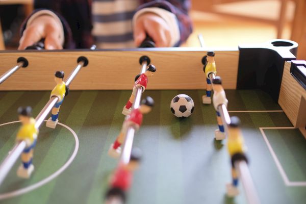 The image shows a foosball table with players in yellow and red uniforms, a ball in the center, and someone playing.