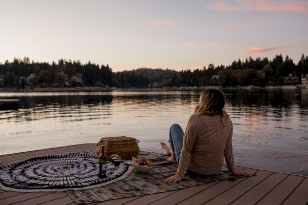 A person sits on a lakeside dock with a picnic setup, enjoying a tranquil sunset view over the water, surrounded by trees.