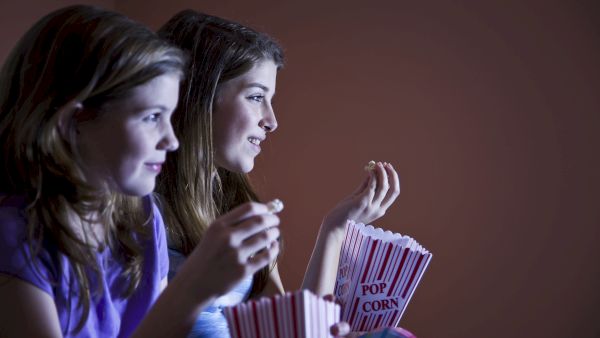 Two people are watching something, holding popcorn boxes, in a dimly lit room.
