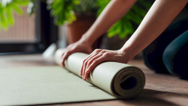 Hands rolling up a yoga mat on a wooden floor, with green plants in the background.