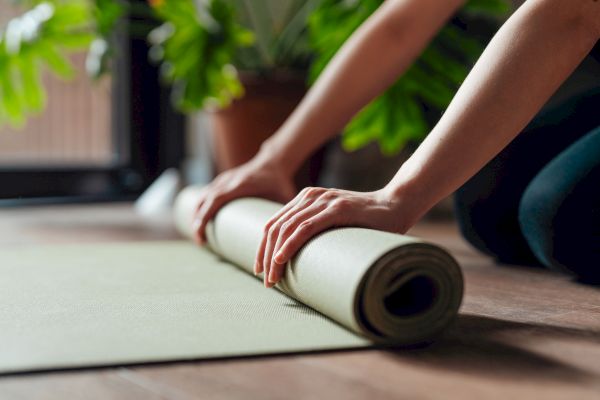 Hands rolling up a yoga mat on a wooden floor, with green plants in the background.