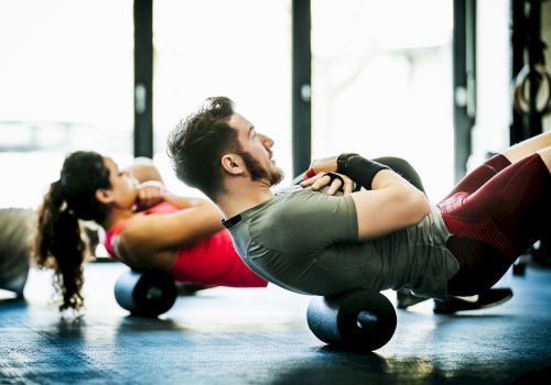 Two people are exercising using foam rollers on the floor, in a brightly lit gym.