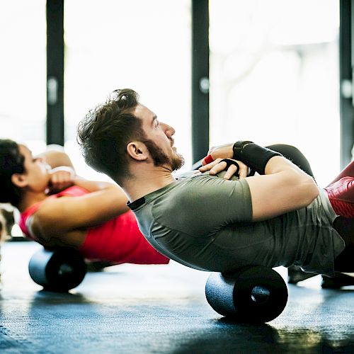 Two people are exercising using foam rollers on the floor, in a brightly lit gym.