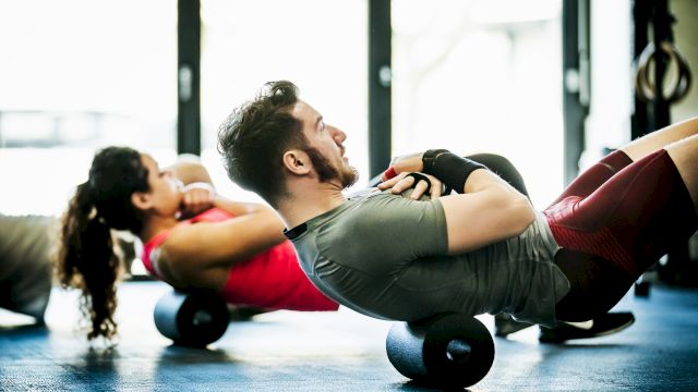 Two people are exercising using foam rollers on the floor, in a brightly lit gym.