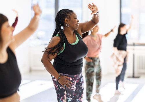 A group of people in a dance class, wearing activewear, engaged in synchronized movement with sunlight streaming through large windows.